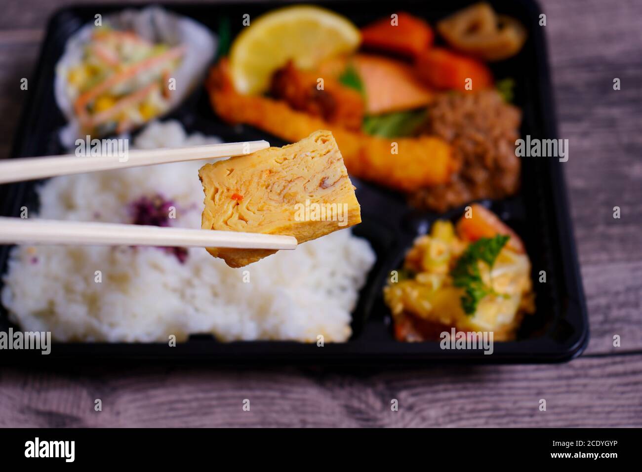 Hölzerne Essstäbchen, die ein saftiges Stück Tofu vor einer verschwommenen japanischen 'Makunouchi' Bento-Box halten. Bento-Boxen sind in Japan sehr beliebt. Stockfoto
