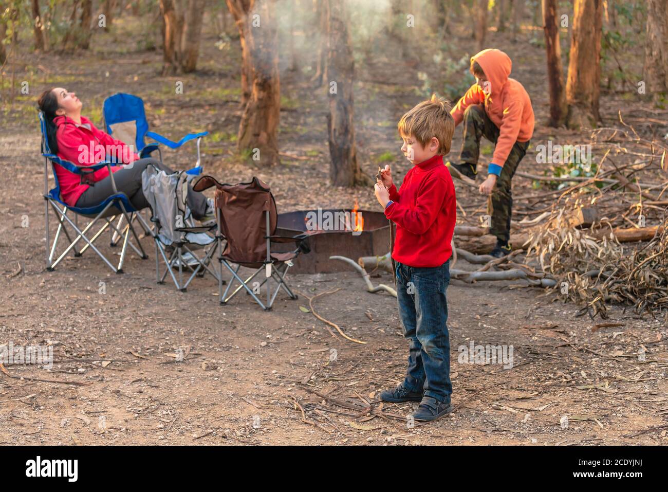 Kinder brennen Feuer auf Kuitpo Wald Campingplatz, während ihre Mutter entspannt in den Schulferien auf dem Stuhl Stockfoto