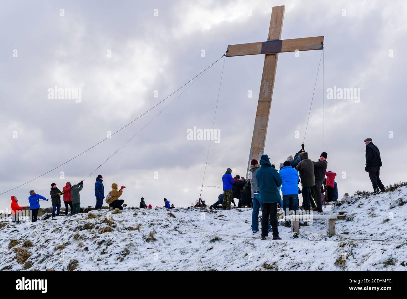 Menschen (Christen) verwenden Seile, um ein riesiges schweres hölzernes Osterkreuz (Faith Symbol) auf einem verschneiten Hügel zu heben - der Chevin, Otley, Yorkshire, Großbritannien. Stockfoto