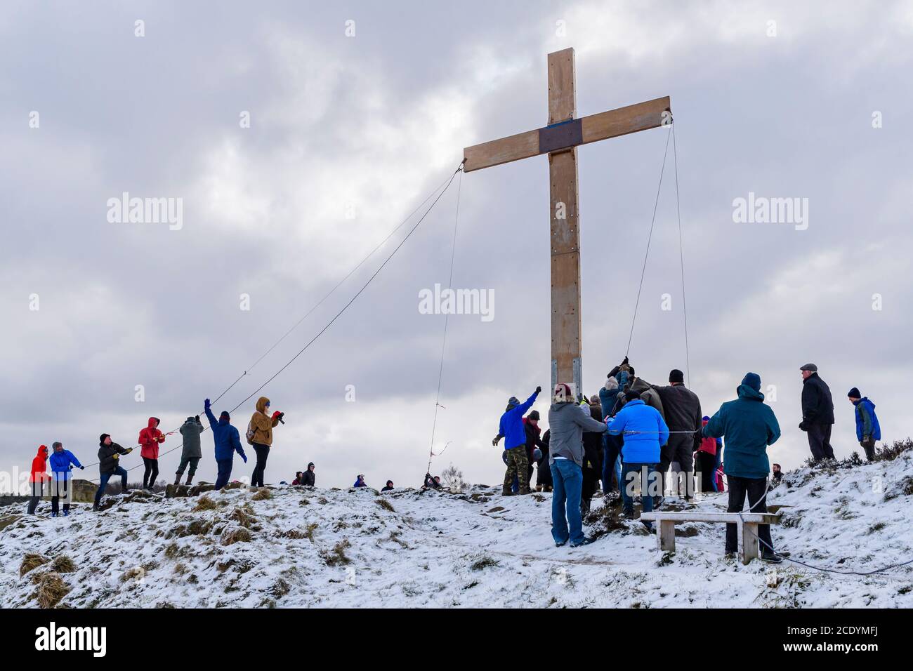 Menschen (Christen) verwenden Seile, um ein riesiges schweres hölzernes Osterkreuz (Faith Symbol) auf einem verschneiten Hügel zu heben - der Chevin, Otley, Yorkshire, Großbritannien. Stockfoto
