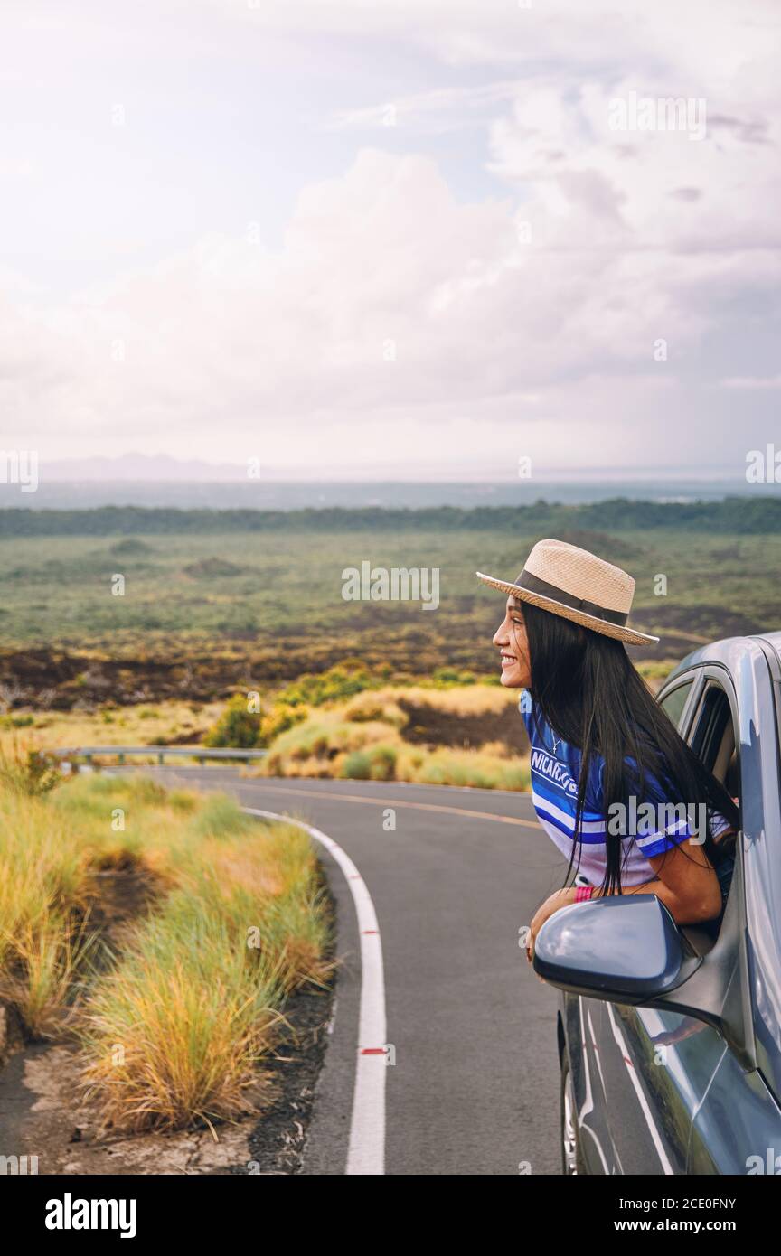 Eine junge Frau, die einen Hut trägt, der sich aus einem lehnt Autofenster mit einer schönen Landschaft im Hintergrund Stockfoto