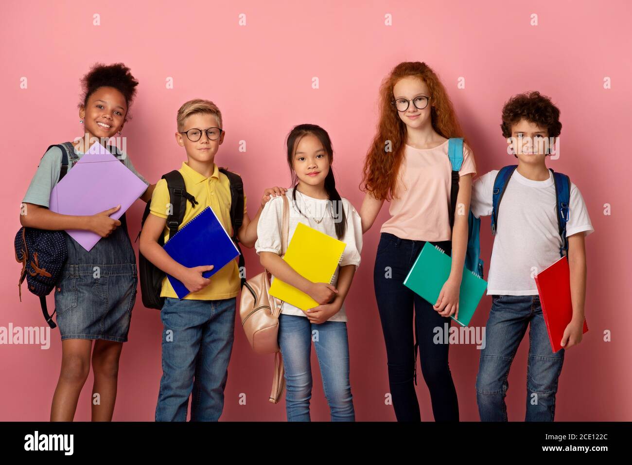 Bereit für die Schule. Kinder verschiedener Nationalitäten mit Notizbüchern und Rucksäcken, die auf die Kamera auf rosa Hintergrund schauen Stockfoto