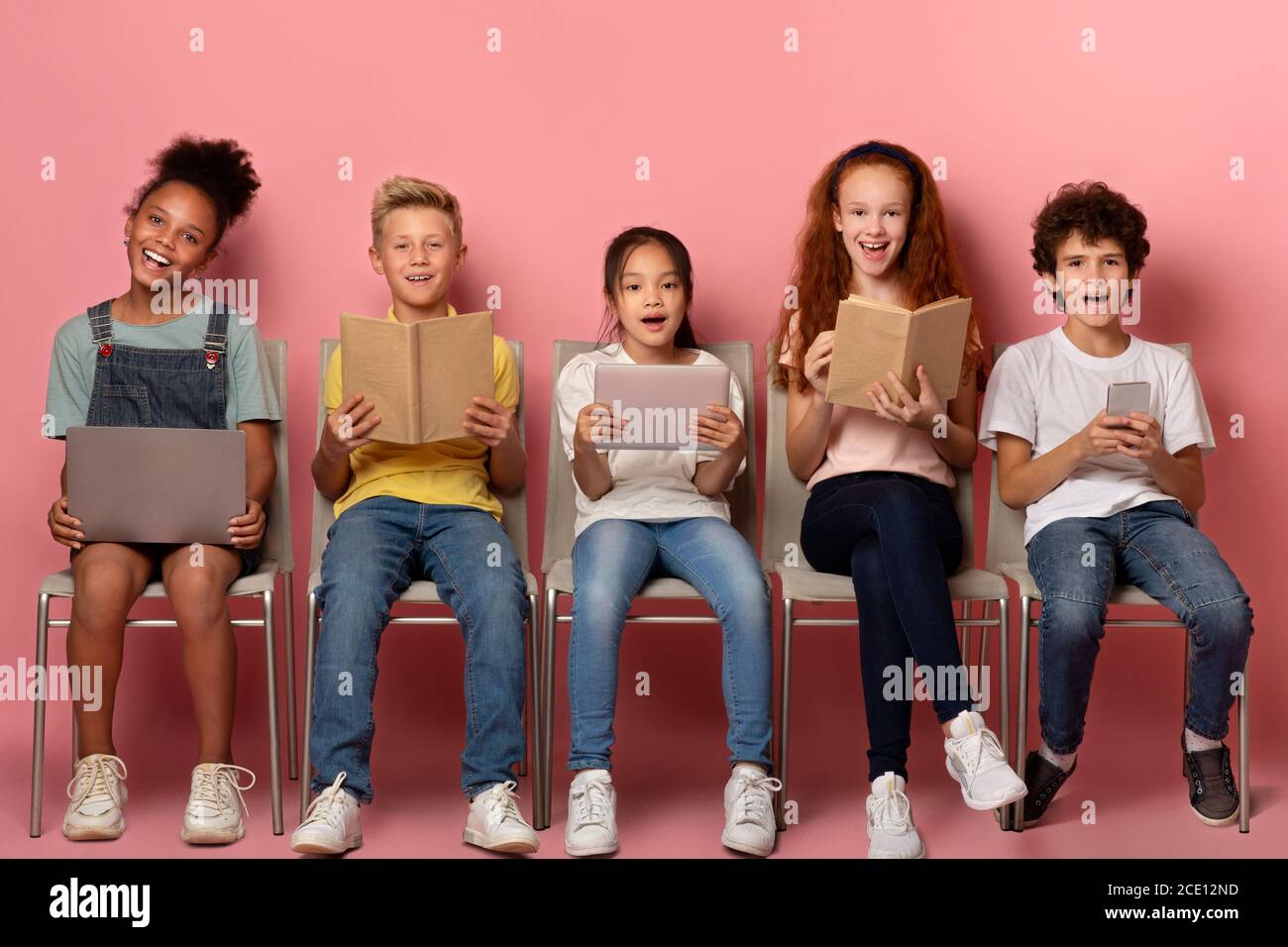 Aufgeregt Kinder mit Büchern und Gadgets für Fernunterricht sitzen Auf Stühlen über pinkem Studiohintergrund Stockfoto