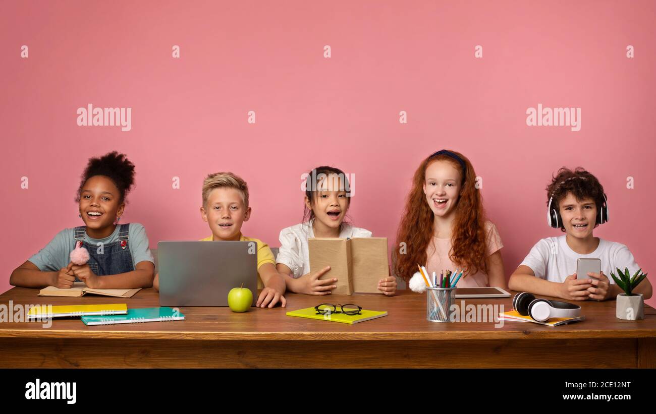 Fernschulbildung. Gruppe von Jungen und Mädchen mit Lernmaterialien und Geräten sitzen am Schreibtisch über rosa Hintergrund Stockfoto