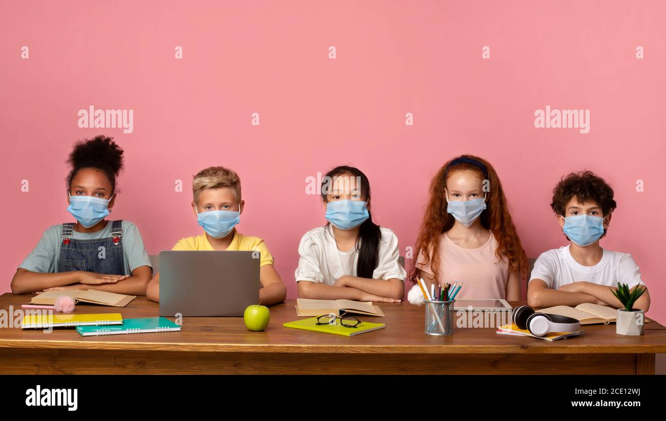 Aufklärung während des Virenausbruchs. Diverse Schulkinder in medizinischen Masken sitzen am Schreibtisch auf rosa Hintergrund, freier Platz Stockfoto