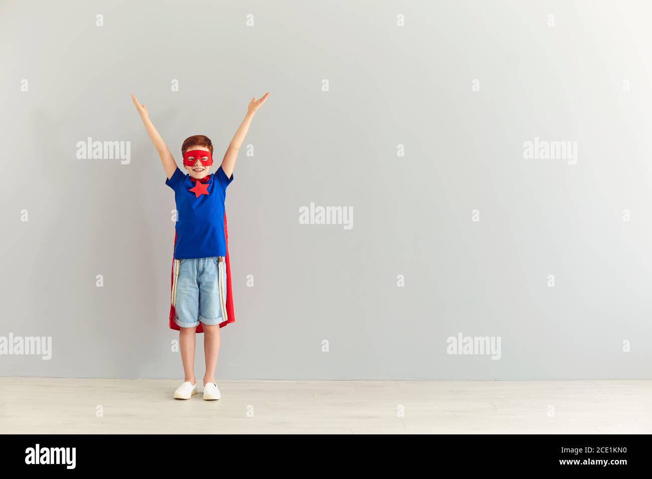 Lächelnder Junge im Kostüm des Superhelden stehend mit erhobenen Händen Auf grauem Hintergrund Stockfoto