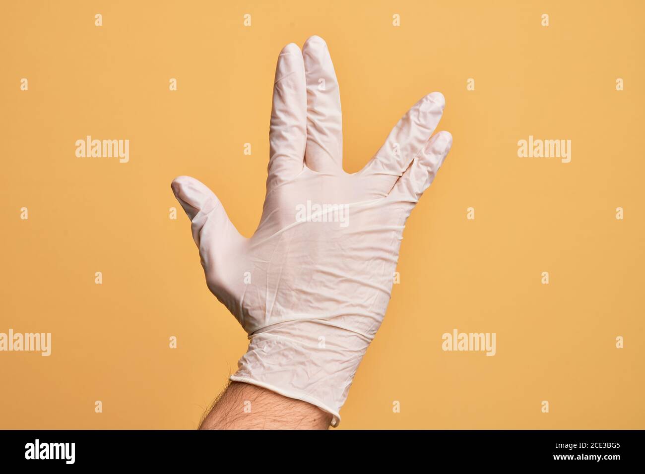 Hand des kaukasischen jungen Mannes mit medizinischen Handschuh über isoliertem gelben Hintergrund Gruß tun Vulcan Salute, zeigt Rückseite der Hand und Finger, fre Stockfoto