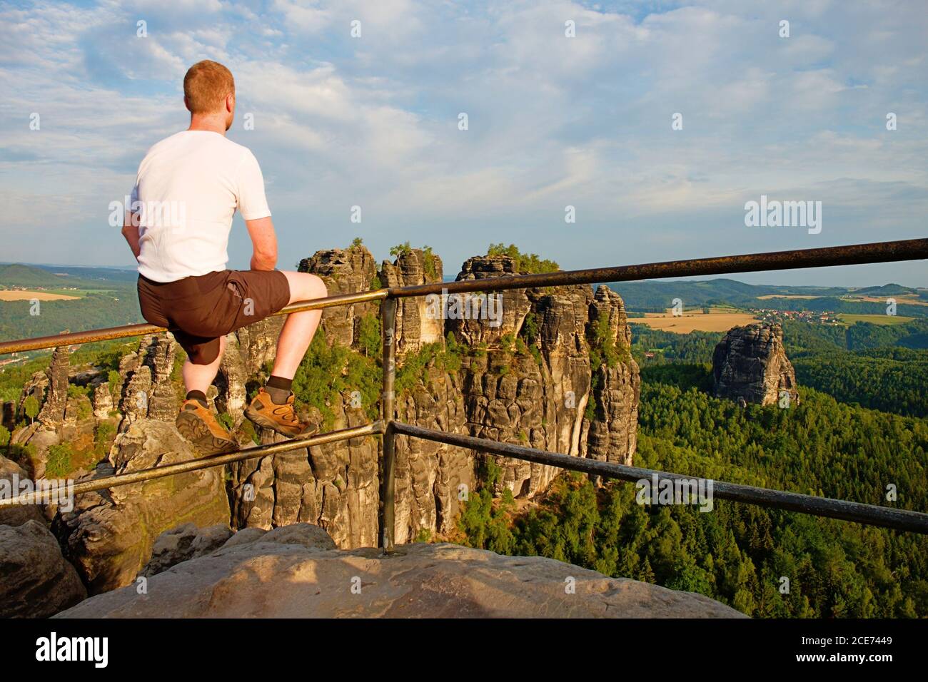 Ingwer Mann sitzen auf dem Geländer auf dem Gipfel des Felsens und beobachten die Landschaft. Sonniger Tag in felsigen Bergen. Wanderer mit grauem Hemd, Hose A Stockfoto