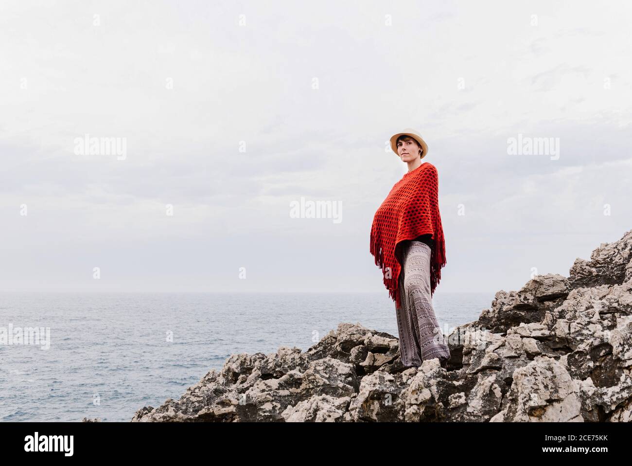 Von unten voller Länge der Frau in stilvoller Kleidung stehen auf dem Rand der felsigen Klippen gegen bewölkten Himmel und bewundern Seeslandschaft während des Urlaubs an der asturischen Küste von Spanien Stockfoto