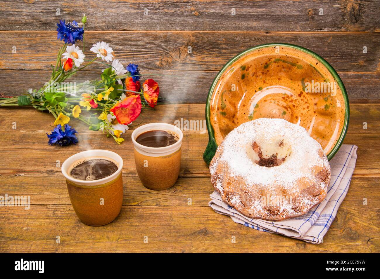 Ein Gugelhupf mit seiner Keramik-Backform und zwei Tassen Kaffee und einem Feld Blumen Geburtstagsstrauß auf einem Holztisch mit Holzhintergrund. Stockfoto