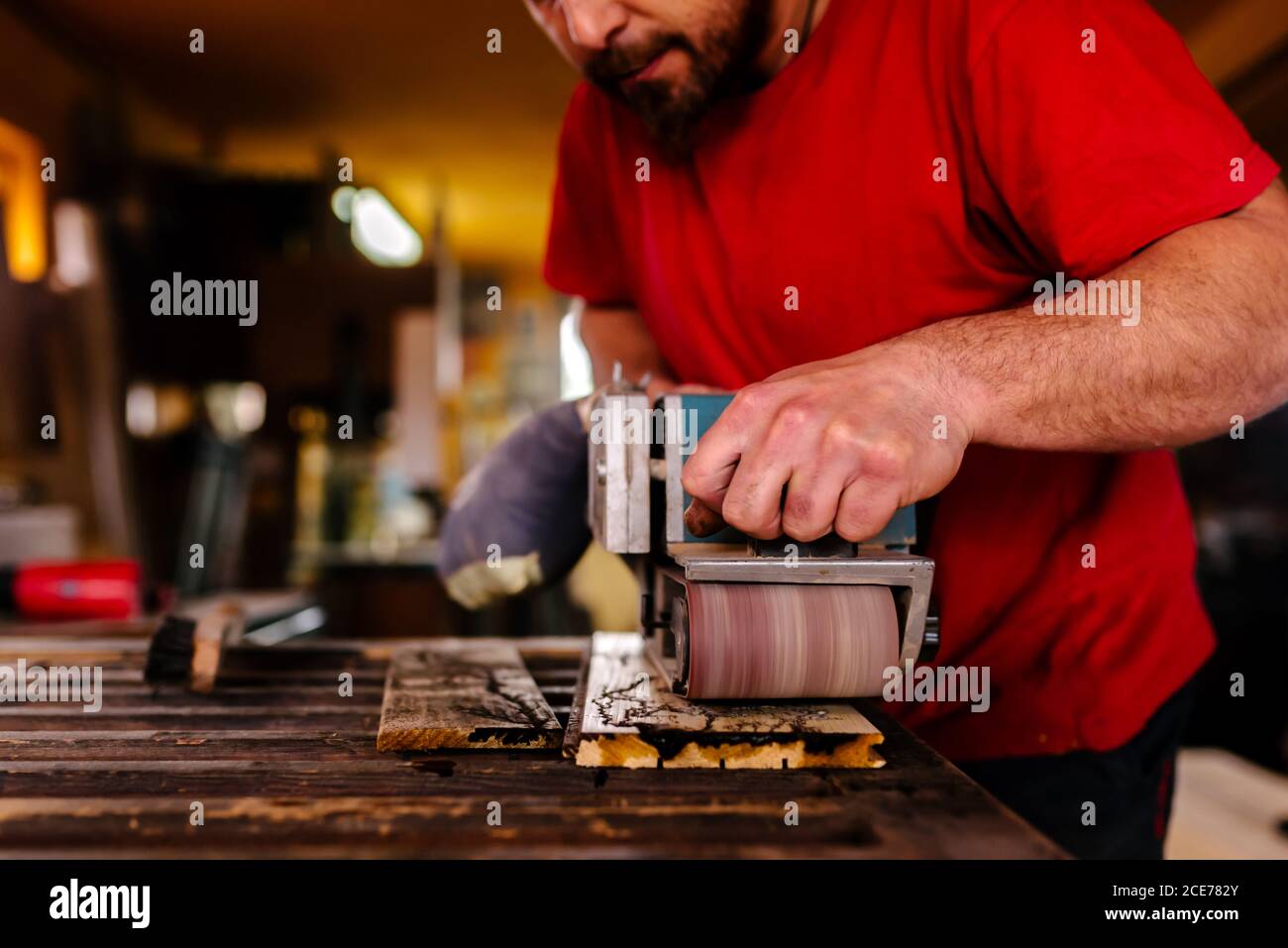 Konzentrierter männlicher Tischler, der an der Werkbank in der Werkstatt und beim Polieren steht Holzbrett mit Bandschleifmaschine Stockfoto