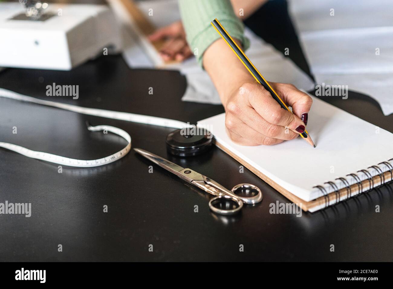 Junge ethnische Näherin in Brillen Notizen stehend nach vorne gelehnt Bei der Verwendung von Lineal am Tisch mit Papier und Schere in Studio im Loft-Stil Stockfoto