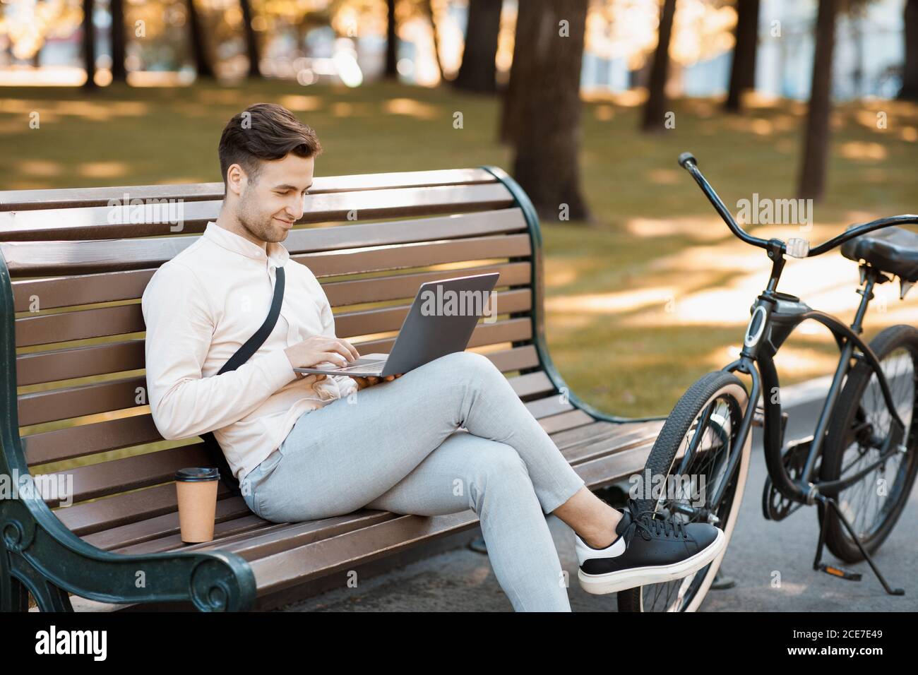 Arbeiten während der Pause im Freien. Attraktiver Kerl sitzt auf der Bank, Typen auf Laptop mit Kaffee und Fahrrad Stockfoto