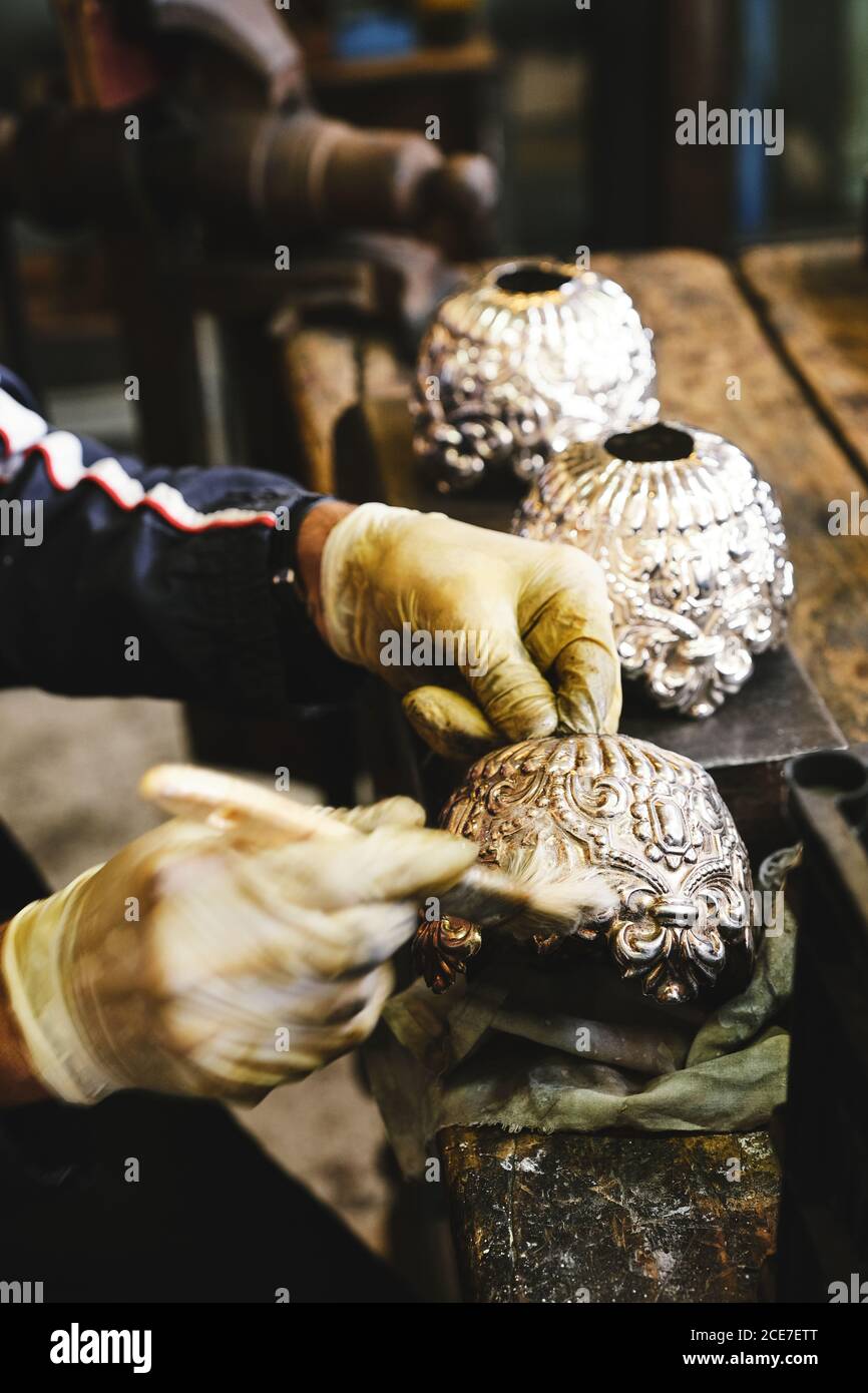 Von oben der Ernte anonymen Handwerker in Handschuhen sitzen bei Alte Holztisch während der Reinigung Gold Vase mit kreativen Ornament Pinsel im Studio verwenden Stockfoto