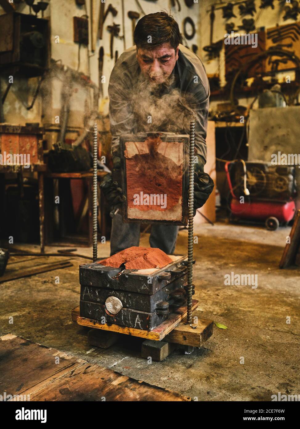 Konzentrierter reifer männlicher Meister, der Musterplatte aus dem Formkasten entfernt Mit Sand bei der Arbeit am Metallguss in der Werkstatt Stockfoto