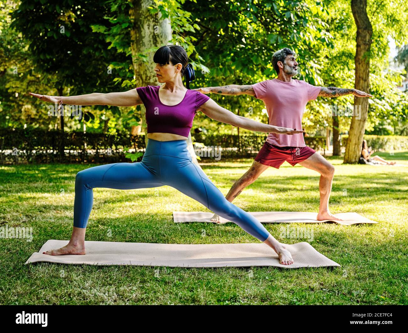 Ruhiges Paar auf Yogamatten, die Yoga in Virabhadrasana machen, während Barfuß im Park an sonnigen Tagen stehen und wegschauen Stockfoto