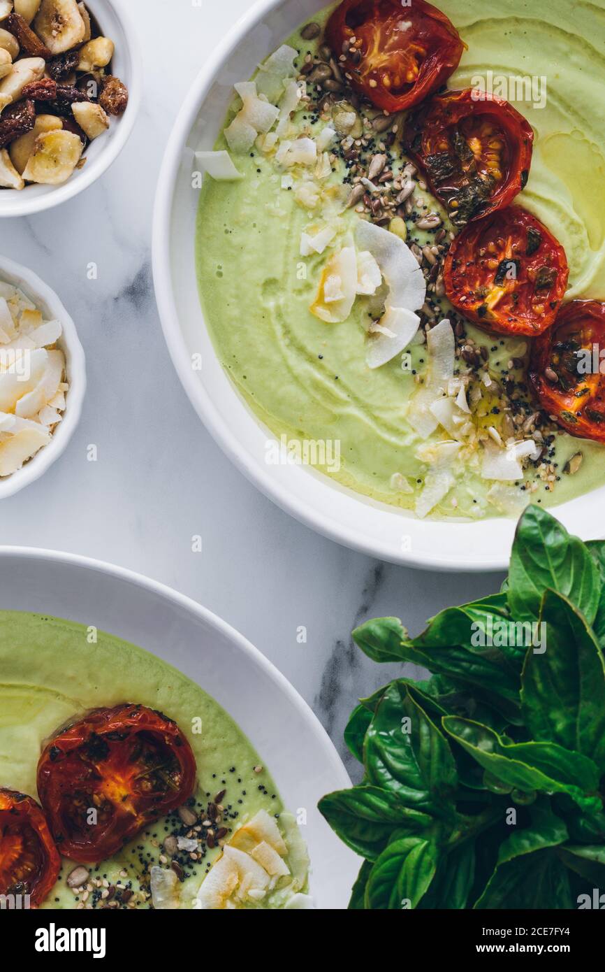 Von oben Schüsseln mit köstlicher Avocado und Apfelsahnesuppe Dekoriert mit sonnengetrockneten Tomaten und gerösteter Kokosraspel Cashewnüsse und frisches Basilikum Stockfoto