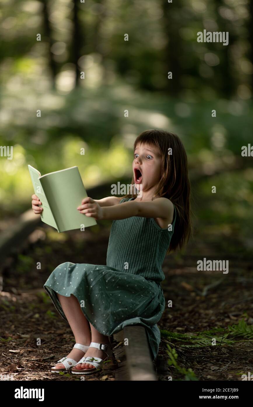Stauned preteen Mädchen im Sommerkleid im Garten sitzen und Demonstration überrascht Gesichtsausdruck beim Lesen interessante Geschichte Stockfoto