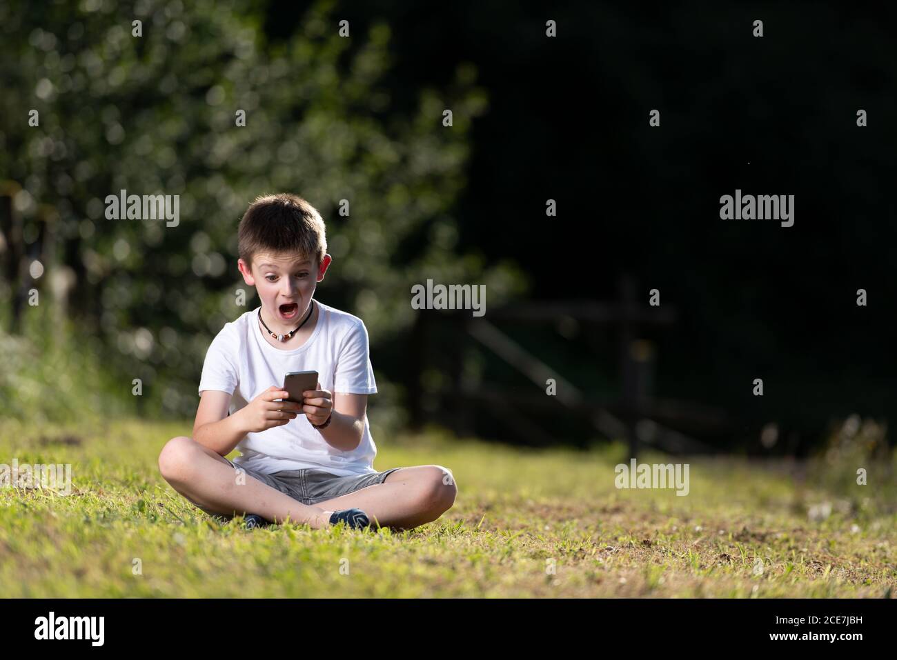 Kind im Sommer tragen sitzen mit gekreuzten Beinen auf grün Rasen und lesen schockierende Nachrichten über Handy Stockfoto