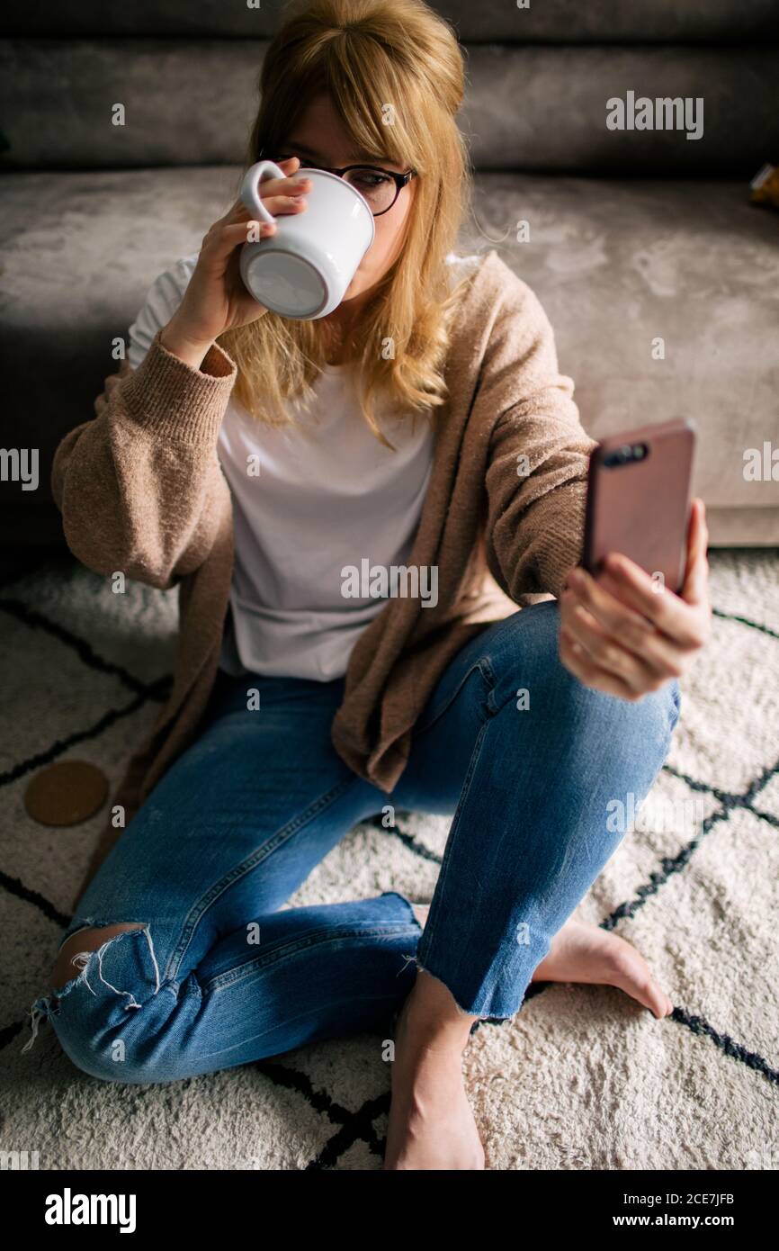 Von oben Inhalt Frau sitzt auf dem Boden auf weichem Teppich Und fotografieren auf dem Handy, während man heißes Getränk trinkt In einem Becher zu Hause entspannen Stockfoto