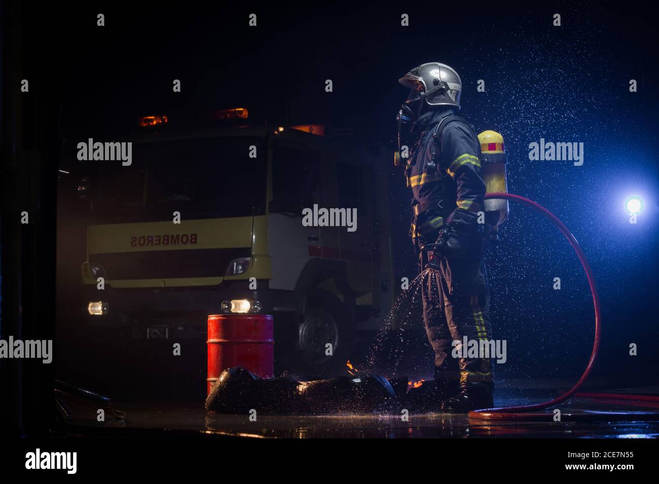 Seitenansicht des Feuerwehrmans mit Schutzuniform und Stehhut Mit Wasserschlauch während regnerischer Nacht in der Nähe von Feuerwehrauto und Wegschauen Stockfoto