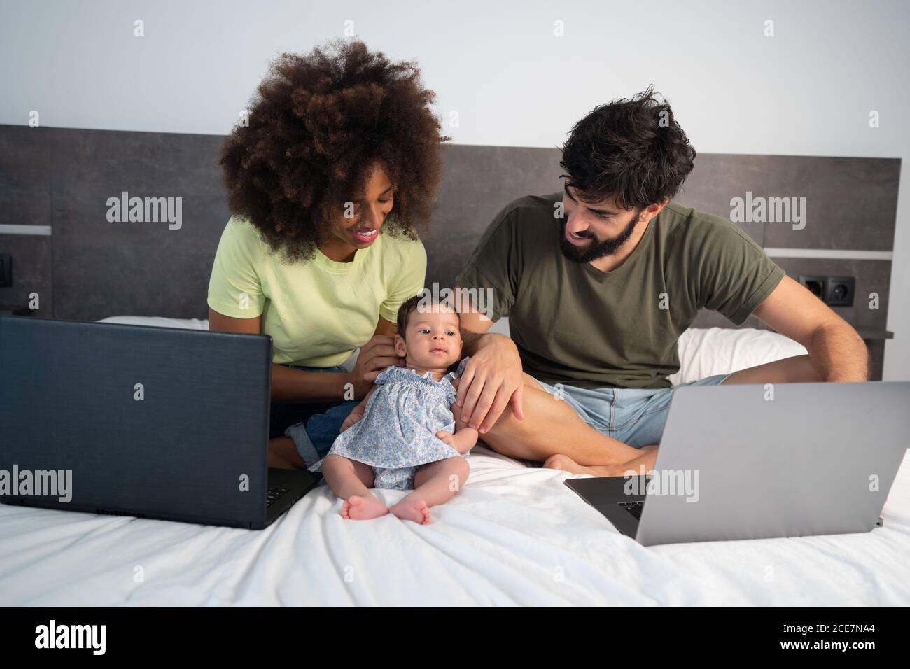 Multiethnisches Paar Freiberufler sitzen auf dem Bett mit niedlichen Kleinkind Tochter und Laptops zu Hause Stockfoto