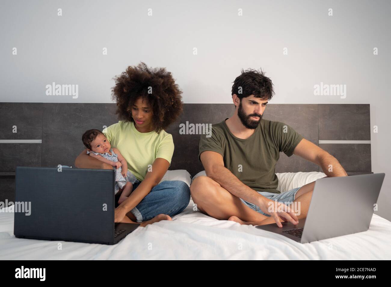 Multiethnisches Paar Freiberufler sitzen auf dem Bett mit niedlichen Kleinkind Tochter und Laptops zu Hause Stockfoto