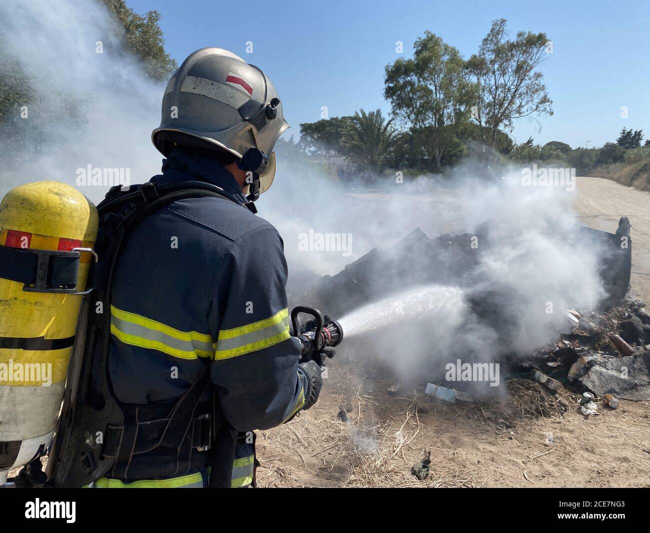 Rückansicht des tapferen Feuerwehrmann in schützender Uniform stehend mit Schlauch und Löschfeuer auf Müllhalde in der Natur Stockfoto