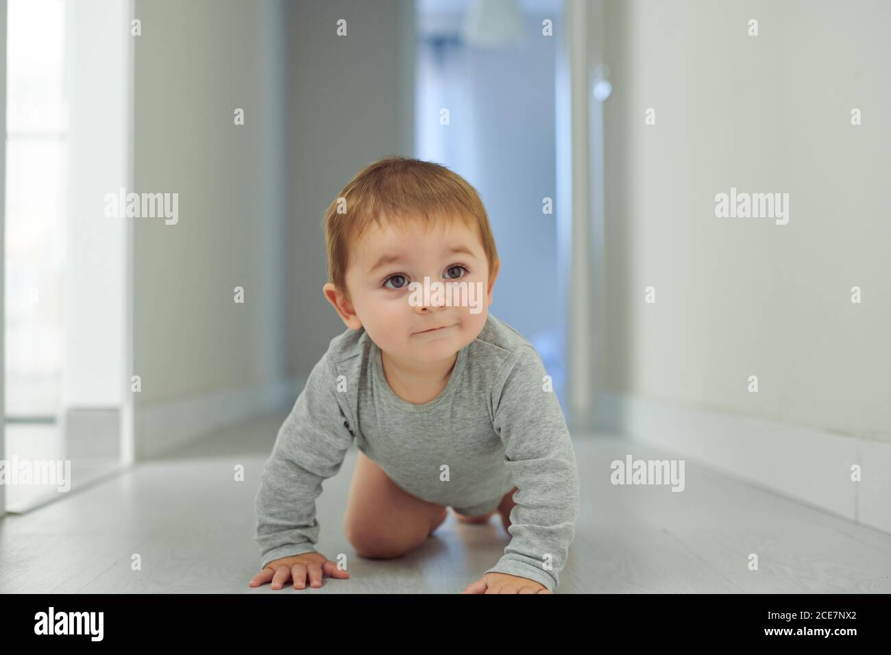 Entzückendes kleines Baby in grauem Body lächelnd beim Krabbeln Boden in gemütlichen Raum zu Hause Stockfoto