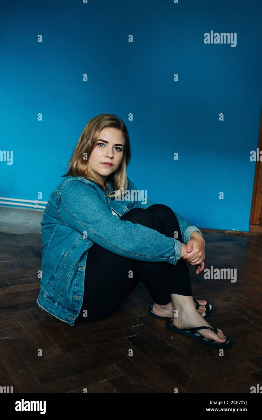 Seitenansicht der jungen Frau in legerer Kleidung umarmen Knie Und auf die Kamera schauen, während man auf dem Boden gegen Blau sitzt Wand Stockfoto