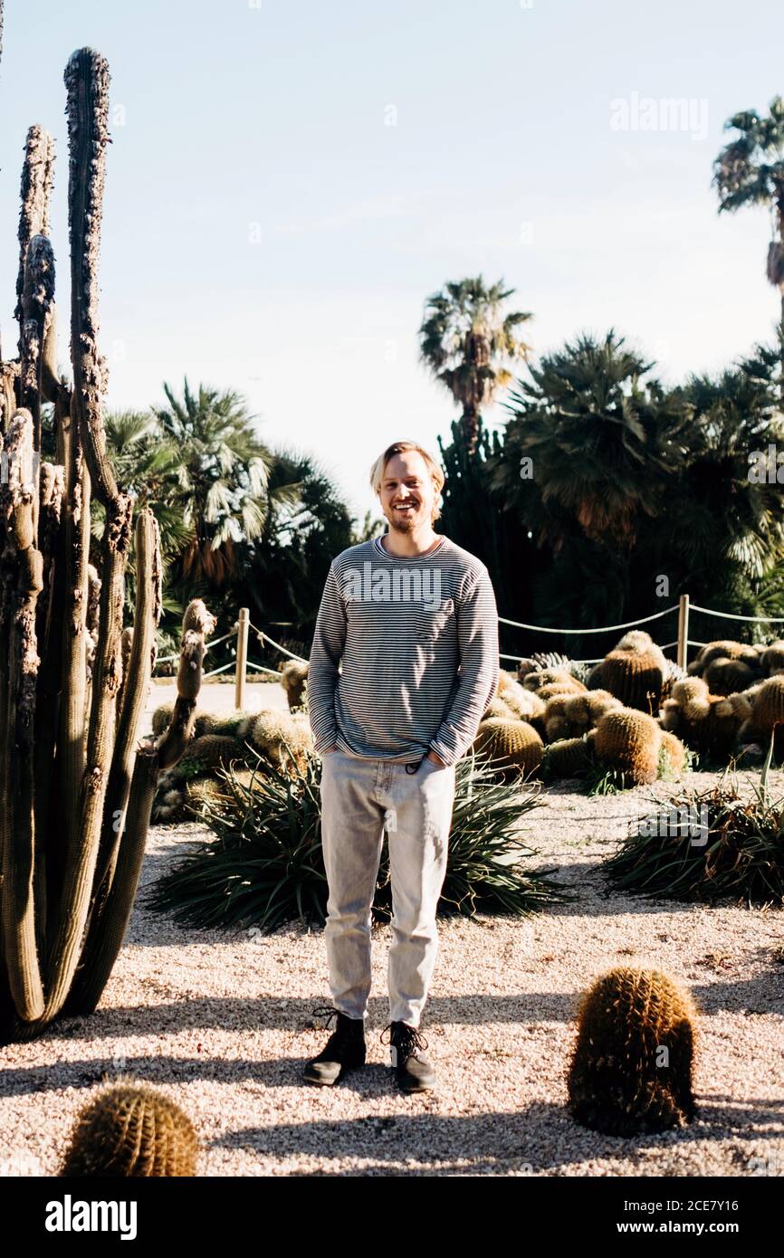 Ganzkörper fröhlich männlich mit Händen in den Taschen lächelnd und Blick auf die Kamera, während in der Nähe von Kakteen an sonnigen Tag im Park in Barcelona, Spanien Stockfoto