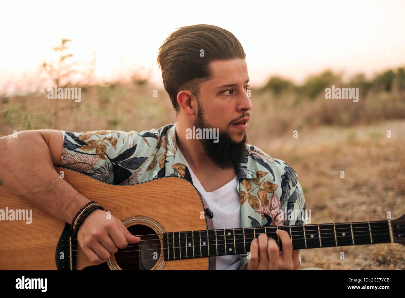 Lächelnder Hipster männlicher Musiker in lässiger Kleidung spielt klassische Gitarre Auf dem Teppich in der Nähe von verblassenem Gras unter heiterem Himmel sitzend Abends Stockfoto