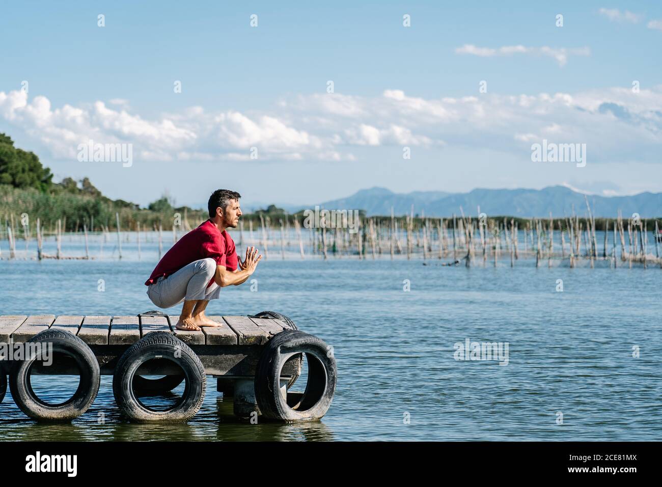 Seitenansicht des konzentrierten Mannes in legerer Kleidung, die die Knöchel streckt Während der Ausübung von Garland Asana während Yoga-Praxis auf hölzernen Pier Stockfoto