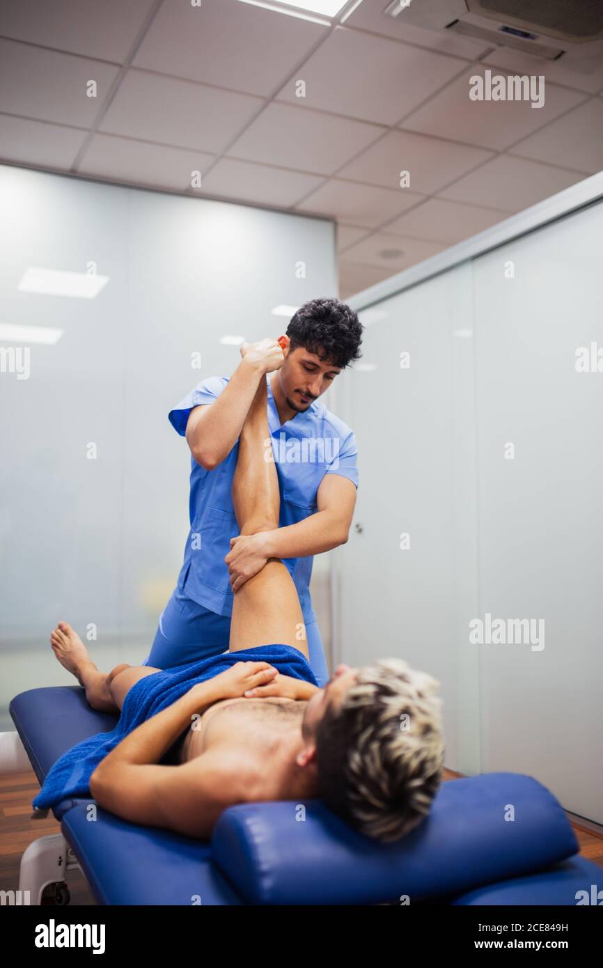 Osteopath in blauer Uniform Untersuchung Knie von unkenntlich schlank männlich Patient mit gefärbten Haaren liegt auf Untersuchungstisch in der Klinik Stockfoto