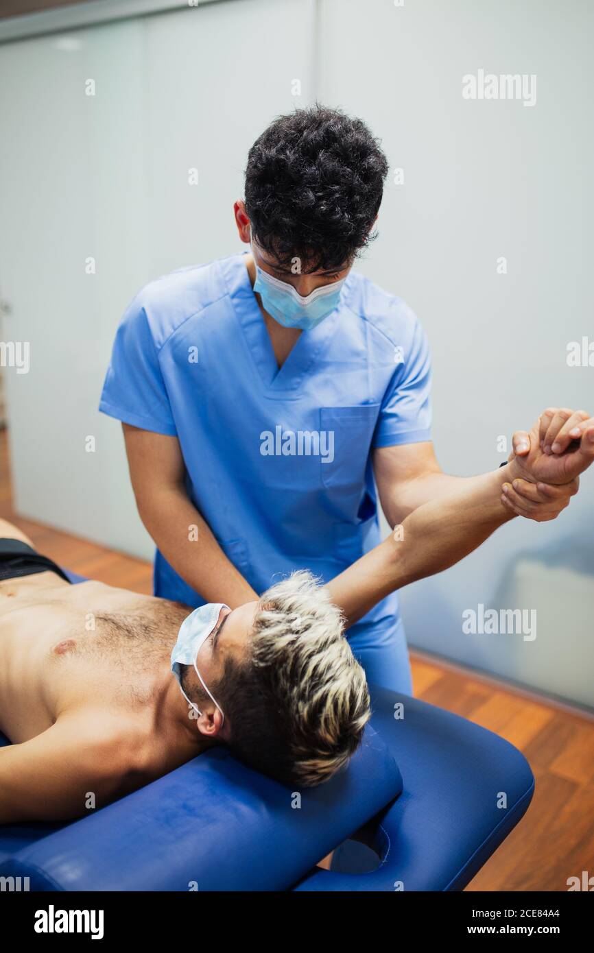 Osteopath mit Maske in blau einheitliche Untersuchungsarm schlank Männlicher Patient mit Maske und gefärbtem Haar liegend auf Untersuchung Tisch in der Klinik Stockfoto