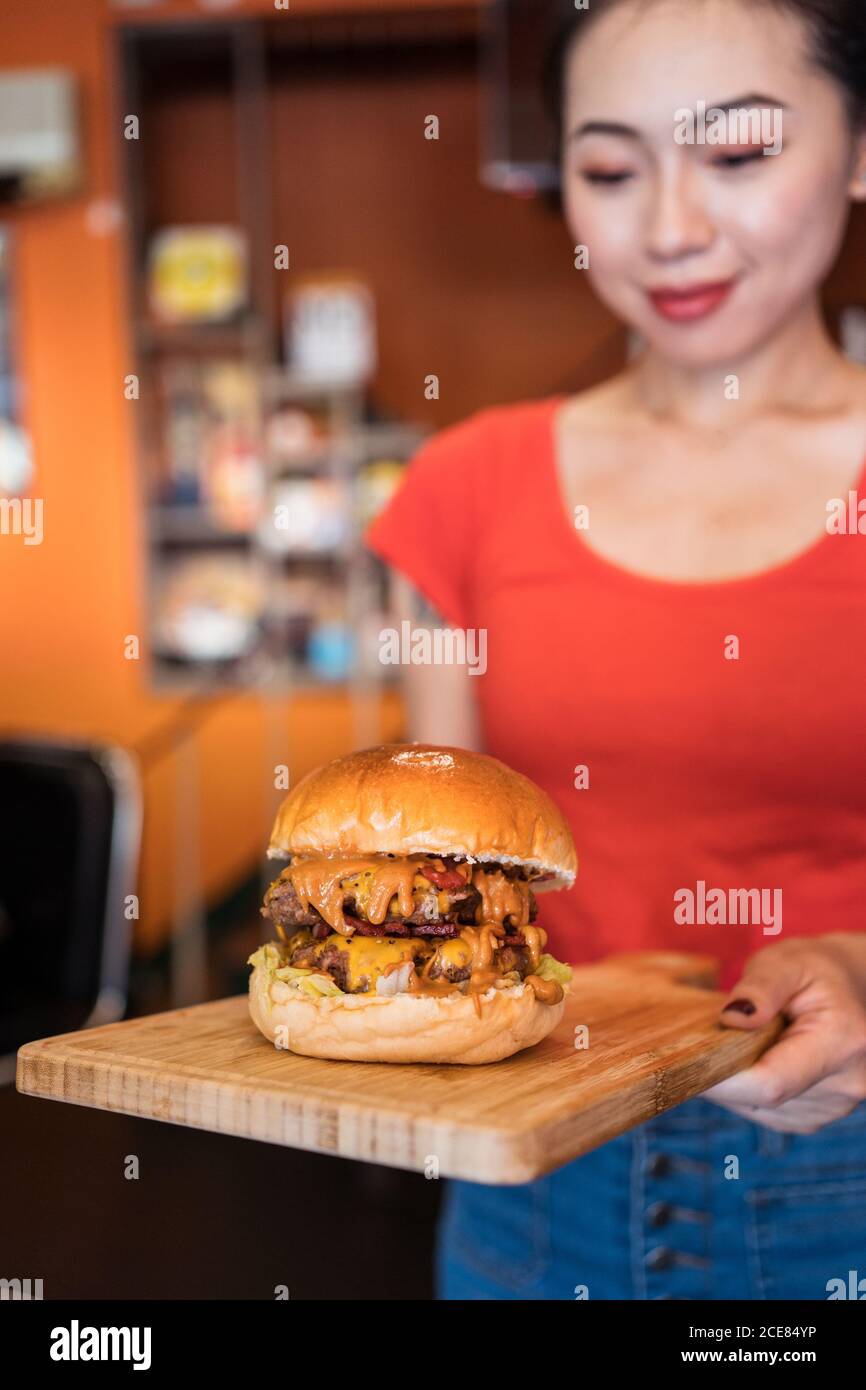Lächelnde ethnische Kellnerin, die Gäste im Café mit Burger versorgt Schneidbrett aus Holz Stockfoto