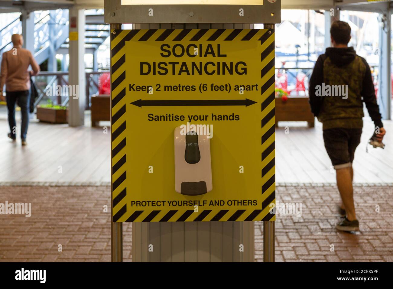 Desinfizieren von Handgel unter einem sozialen Distanzschild am Eingang eines Einkaufszentrums mit Leuten, die vorbeigehen Stockfoto