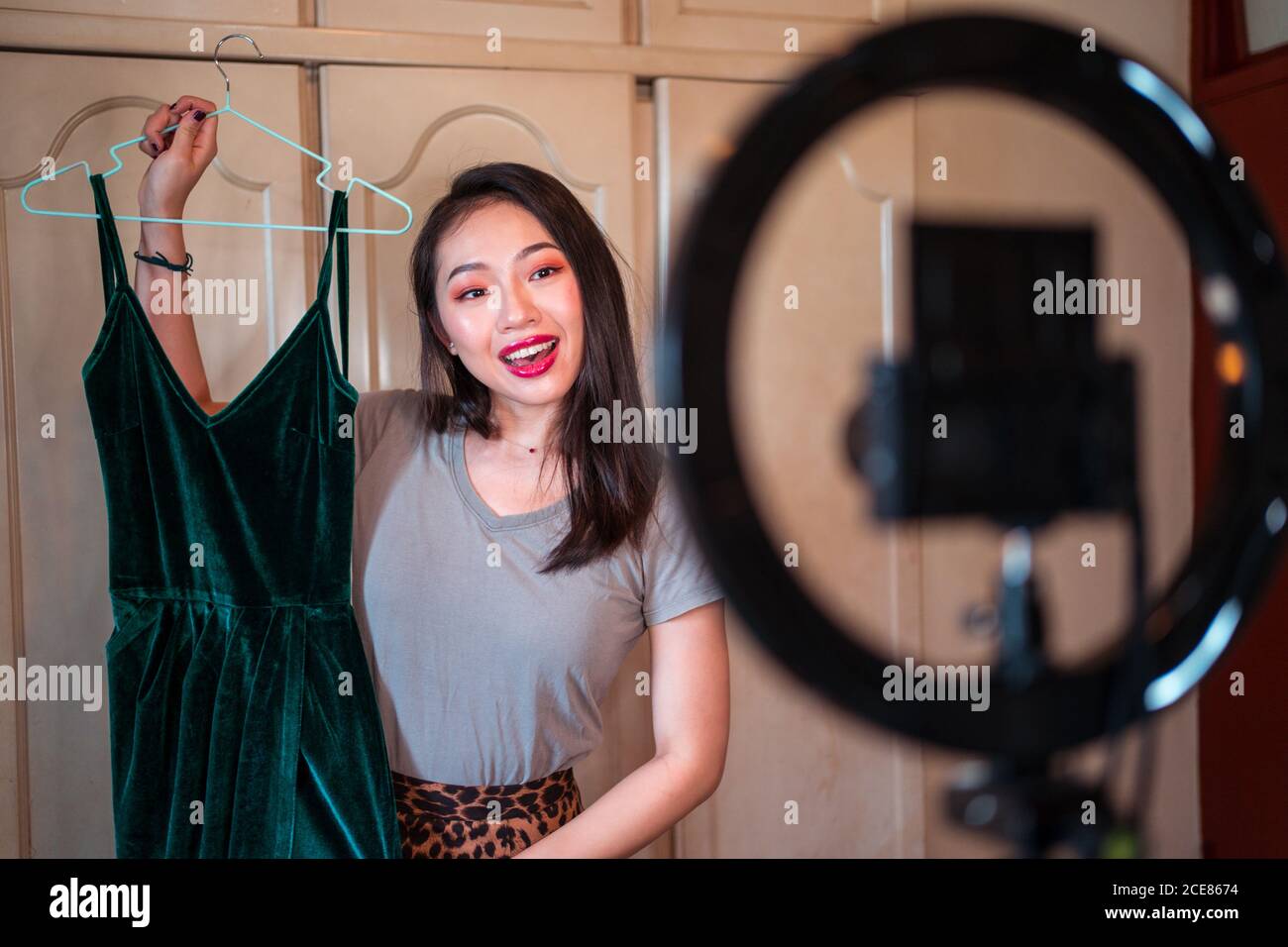 Positive junge asiatische Frau zeigt stilvolle Kleider und geben Beratung Während der Videoaufnahme für Mode-Vlog, während Sie vor stehen Von Stativ mit Kamera und Ringlampe zu Hause Stockfoto