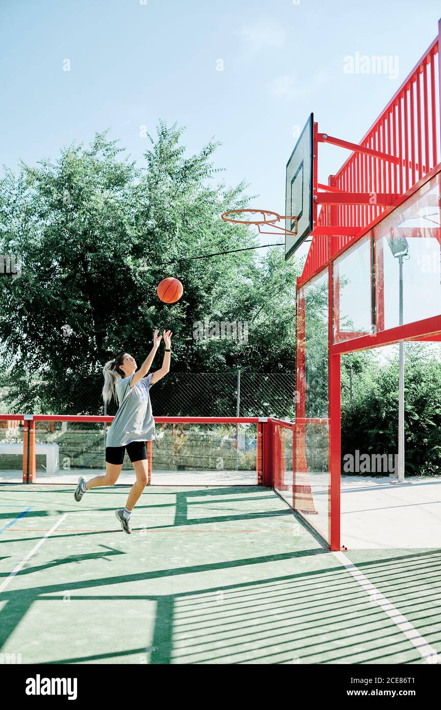 Ganzkörper-Seitenansicht einer jungen, sportlichen Frau, die einen Jump Shot ausführt, während sie allein auf dem Sportplatz Basketball spielt Stockfoto