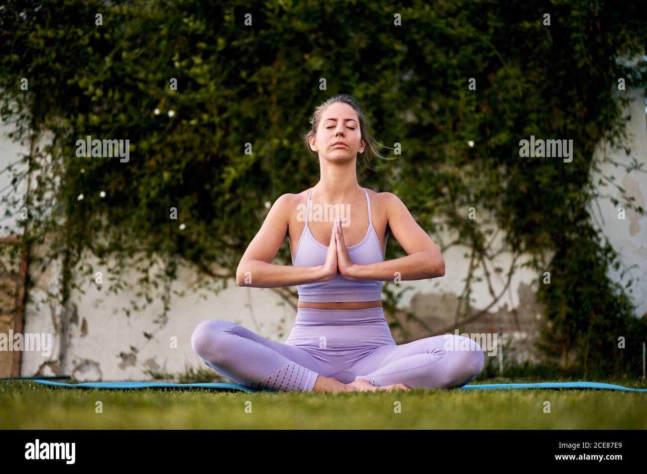 Friedliche Frau in aktiver Kleidung sitzt in Padmasana mit Namaste Geste beim Meditieren und Yoga üben auf Rasen Stockfoto