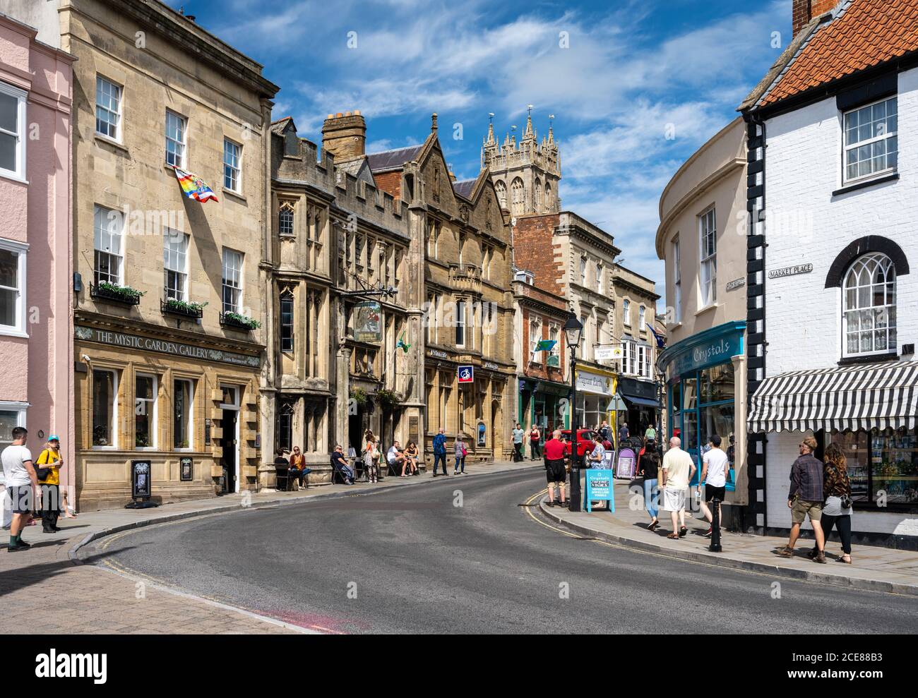 Fußgänger gehen an Geschäften, Pubs und Cafés in den alten Gebäuden der High Street und Market Place im Zentrum der englischen Stadt Glastonbury vorbei. Stockfoto