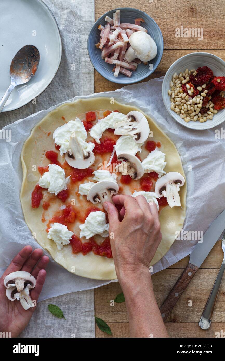 Draufsicht auf Ernte unkenntlich Person Kochen Quiche mit dünn Blätterteig mit Tomatensauce und Pilzscheiben bedeckt Mit frischem italienischen Burrata-Käse in der Nähe Schüssel mit Prosciutto Stockfoto