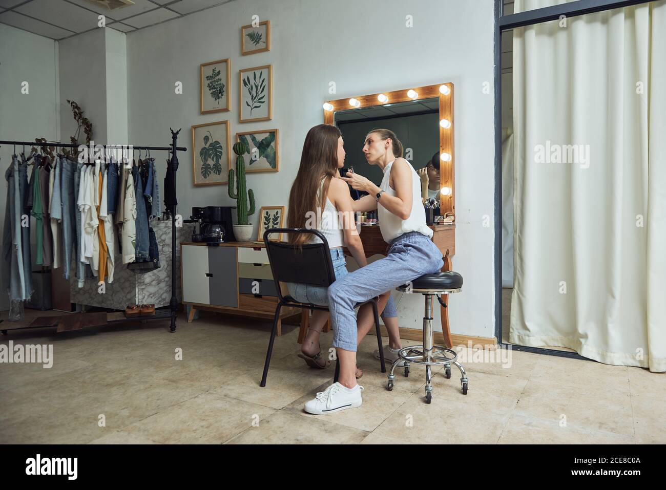 Positive junge weibliche Make-up Künstler tun Kunden Gesicht Make-up mit Pinsel neben gemütlichen großen Spiegel mit Leuchten in zeitgenössischer Schönheit Salon Stockfoto