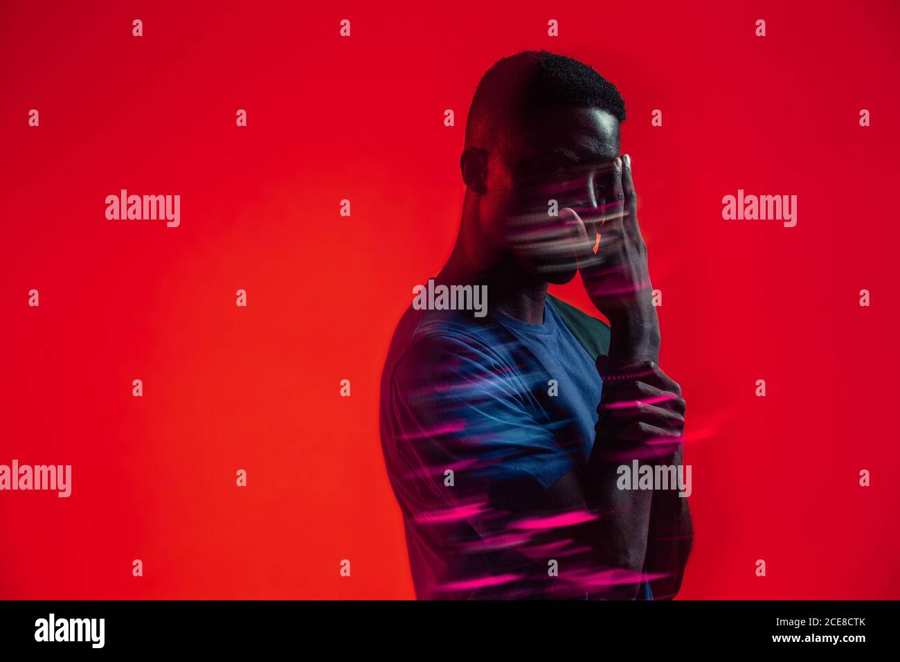 Serious zuversichtlich jungen afroamerikanischen Mann mit bunten Neonlicht Auf Gesicht Blick auf die Kamera vor rotem Hintergrund Stockfoto