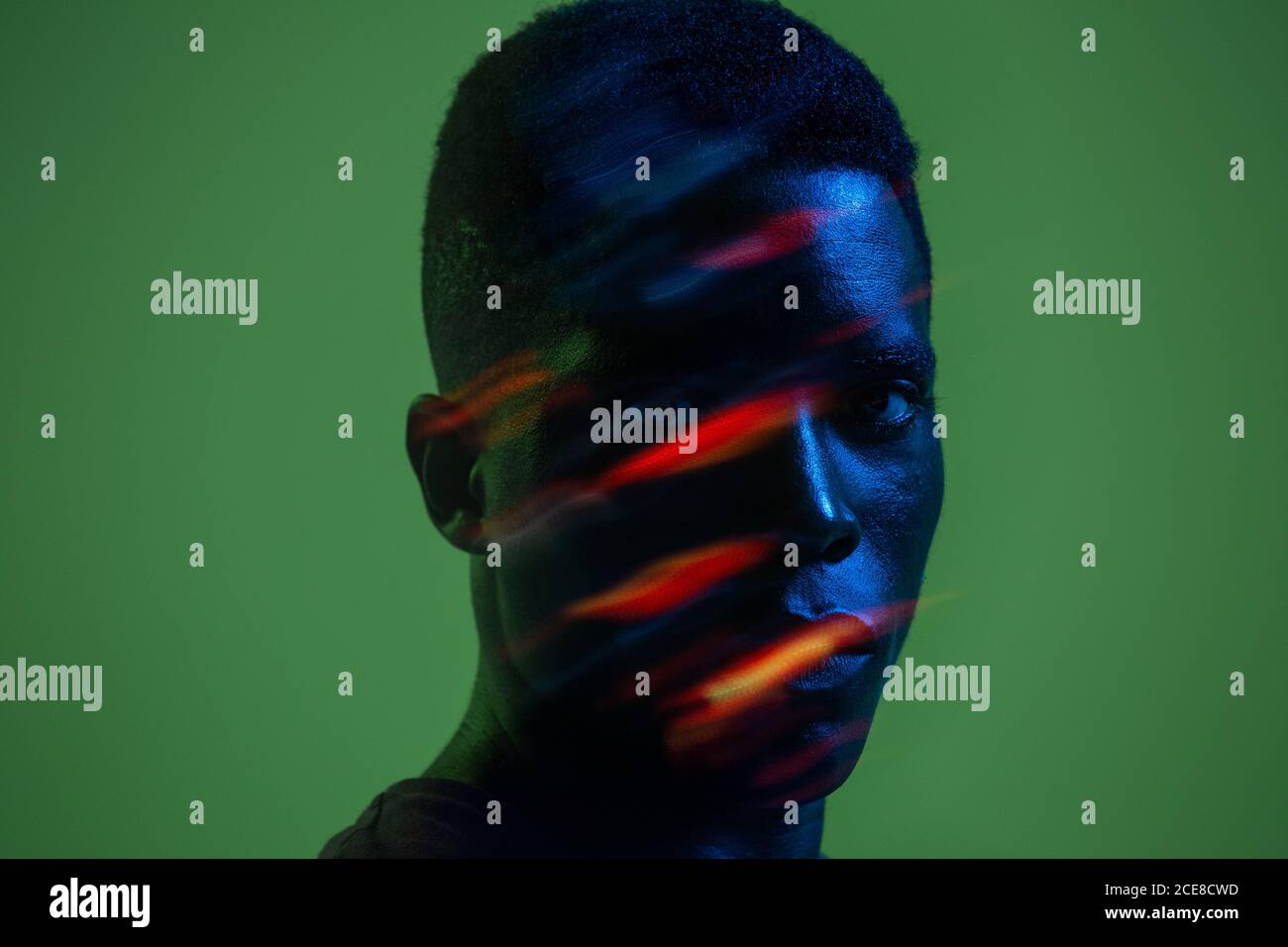 Serious zuversichtlich jungen afroamerikanischen Mann mit bunten Neonlicht Auf dem Gesicht Blick auf die Kamera vor grünem Hintergrund Stockfoto