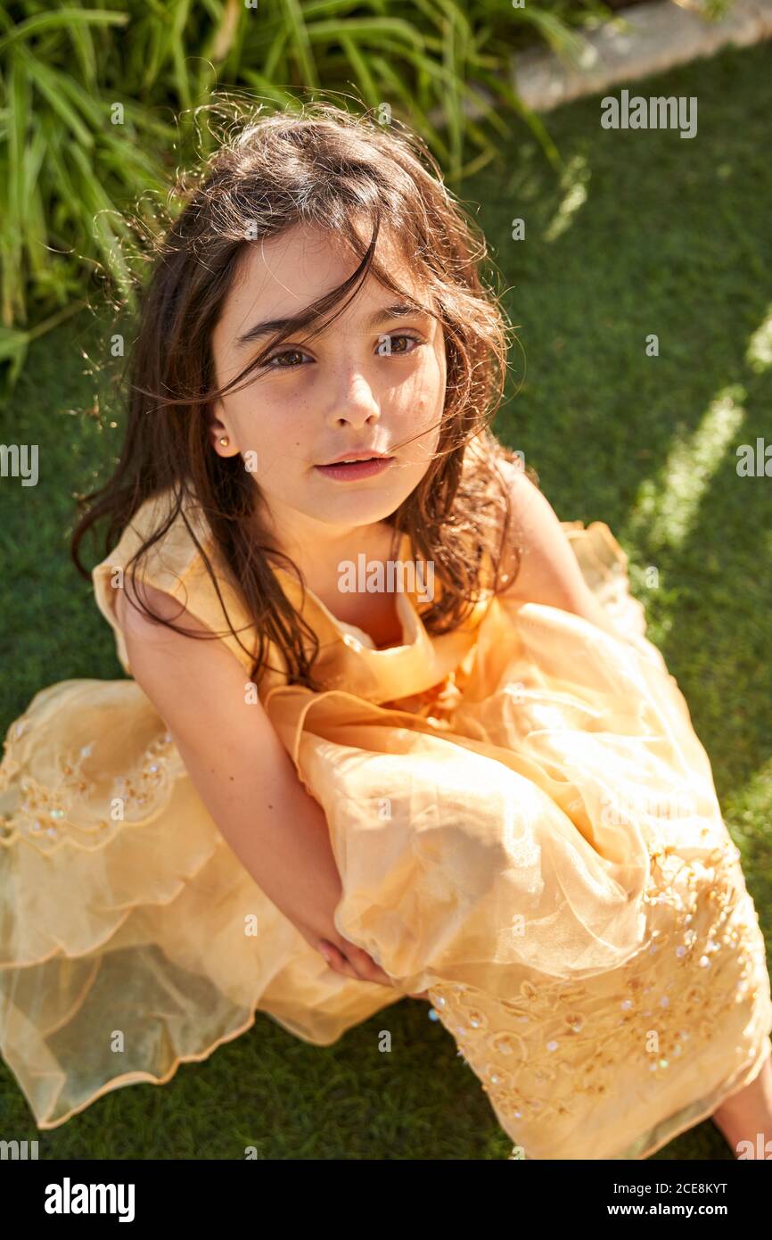 Von oben entzückende ruhige kleine Mädchen mit langen wellig Dunkle Haare  tragen gelbe Spitze Kleid sitzt auf grünem Gras Und Blick auf die Kamera  während der Ruhe im Garten im Sommer Tag