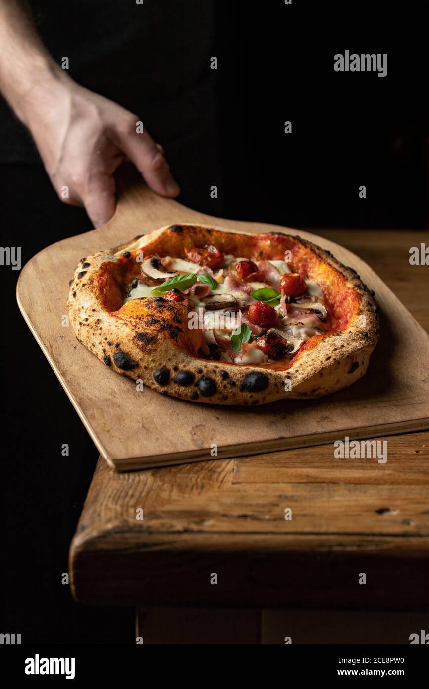 Anonymer Bäcker mit köstlicher Pizza mit Käse und Gemüse platziert Auf Holzschale auf schwarzem Hintergrund Stockfoto