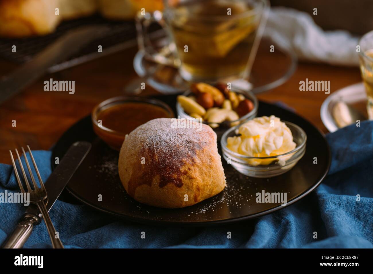 Von oben Keramikplatte mit frischem Brötchen und klein Schüsseln mit Mischung von Nüssen in der Nähe von Marmelade und Butter schließen Zu einer Tasse grünem Tee auf einem zerknitterten Handtuch Stockfoto