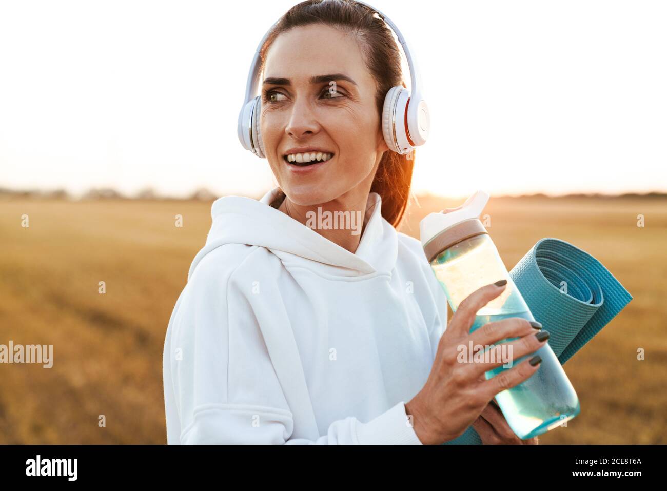 Bild von lächelnd schöne Sportlerin in Kopfhörer Trinkwasser während Yogamatte am goldenen Feld halten Stockfoto