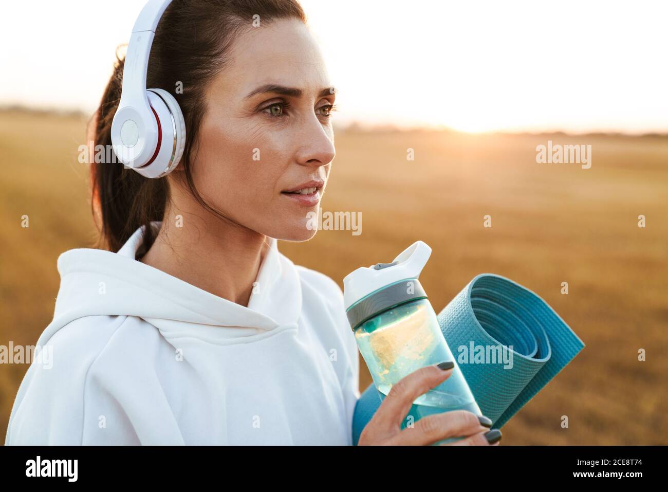 Bild von schönen Sportlerin in Kopfhörer Trinkwasser während Yogamatte am goldenen Feld halten Stockfoto